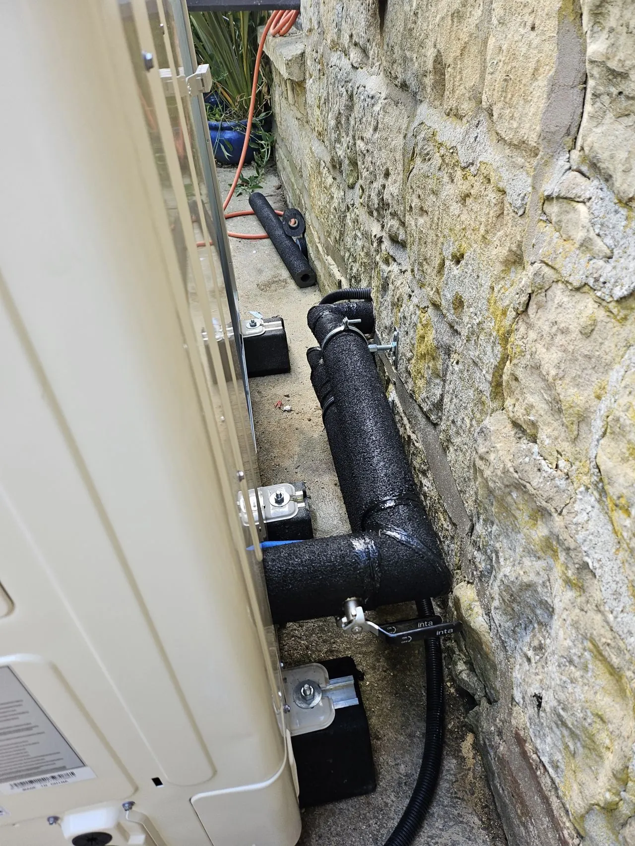 Insulated heat pump pipework routed beside a stone wall.
